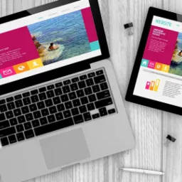 Laptop, tablet i smartfon wyświetlające responsywny projekt strony internetowej z motywem plaży i osoby pływającej w morzu, leżące na drewnianym biurku z akcesoriami biurowymi.