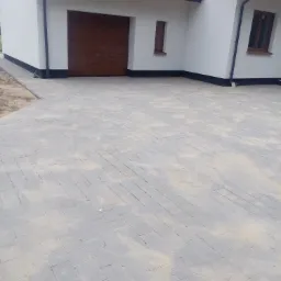 Ułożona szara kostka brukowa na podjeździe przed domem z brązową bramą garażową i oknami z brązowymi ramami.