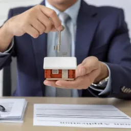 Mężczyzna w garniturze trzyma klucz nad modelem domu z czerwonego drewna z białym dachem, na biurku leżą dokumenty z napisem 'Loan Application'.