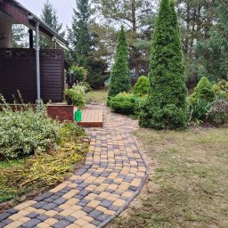 Bartek Clean - Ułożona ścieżka z kostki brukowej w dwóch kolorach prowadząca do domu, otoczona zielenią i drzewami iglastymi. Widok na fragment elewacji domu z drewnianą balustradą.
