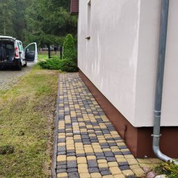 Bartek Clean - Ułożona kostka brukowa w szachownicę przy budynku w Kostrzynie nad Odrą. Widoczny fragment elewacji i rynna spustowa. W tle zieleń i zaparkowany samochód.