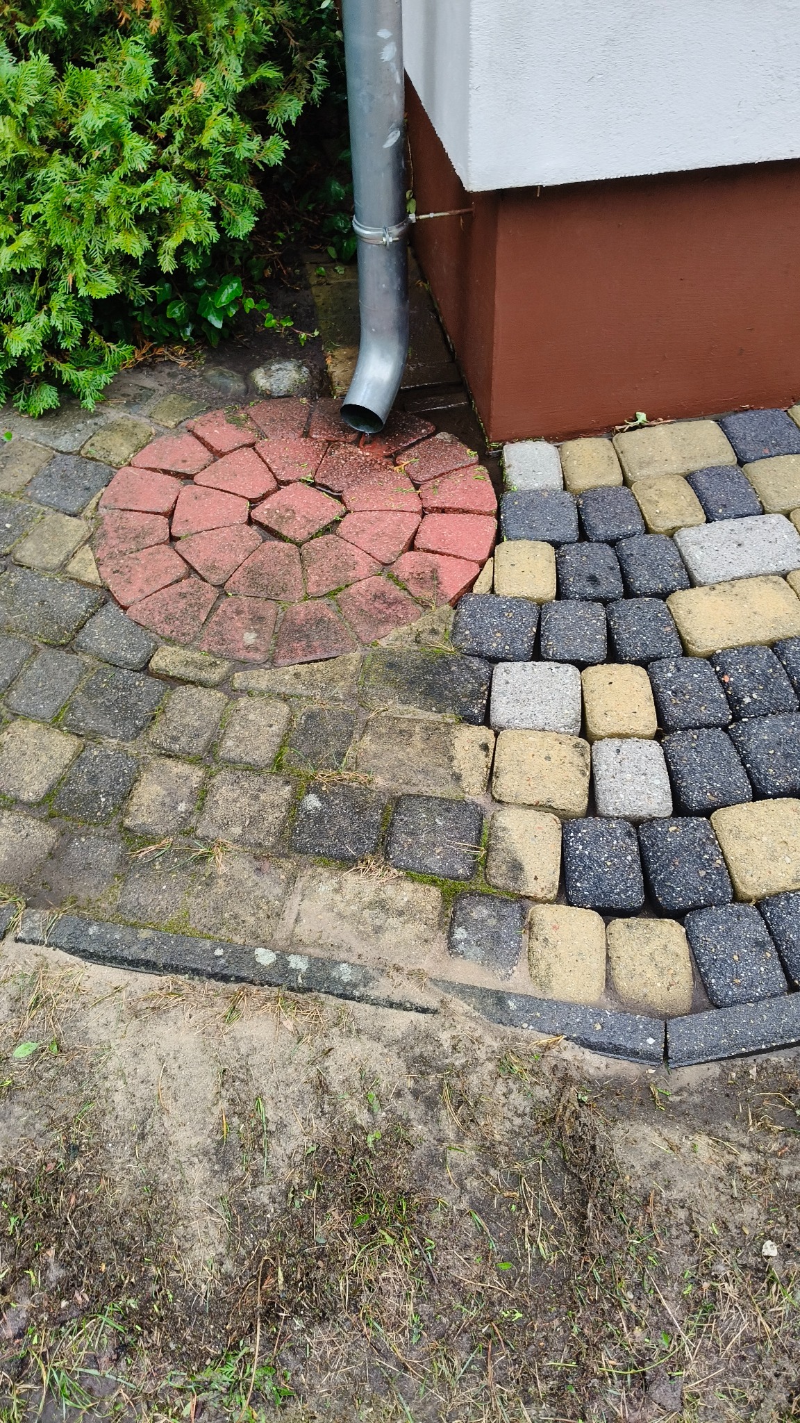 Zanieczyszczona kostka brukowa w różnych kolorach, z widocznym odpływem rynnowym i roślinnością. Część kostki pokryta błotem i mchem, co sugeruje potrzebę czyszczenia.