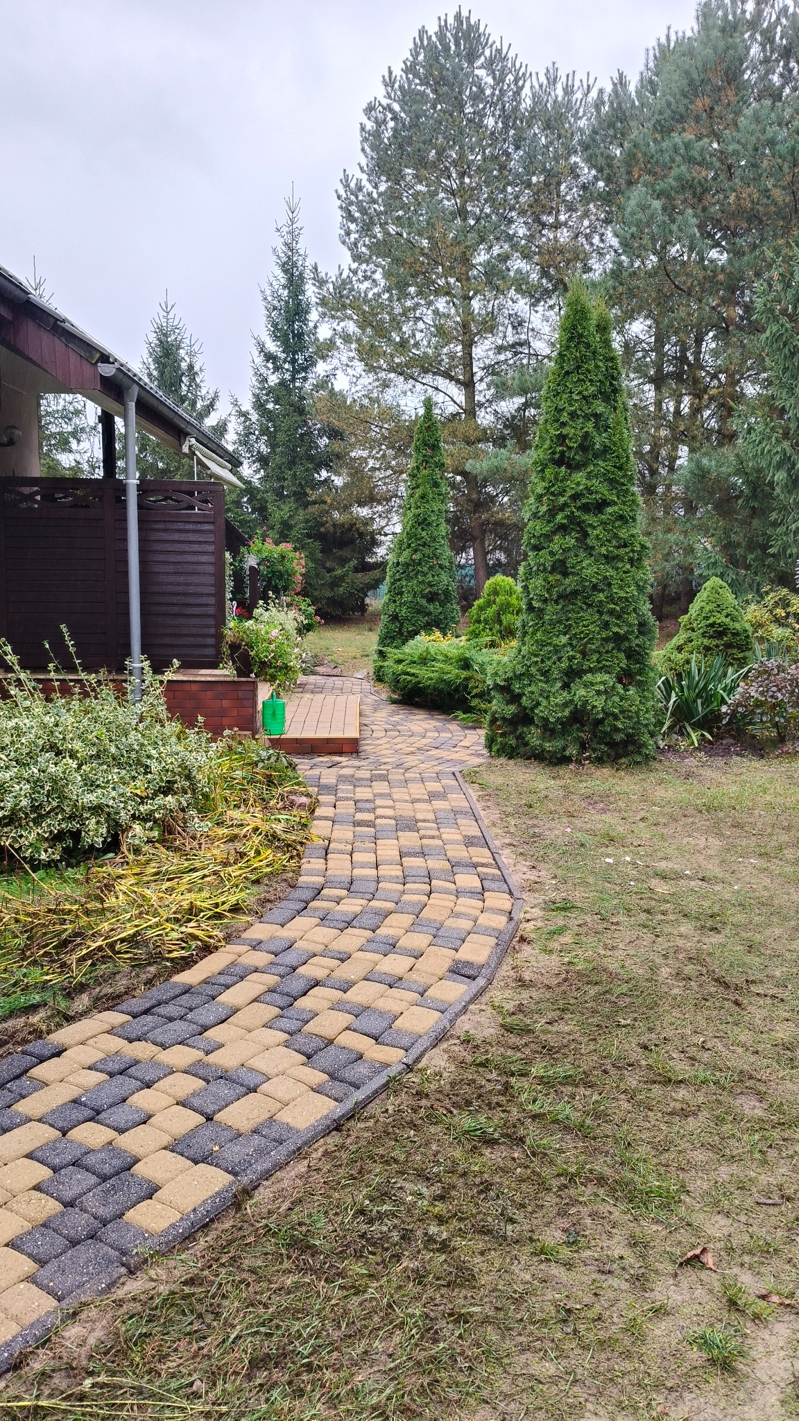Ułożona ścieżka z kostki brukowej w dwóch kolorach prowadząca do domu, otoczona zielenią i drzewami iglastymi. Widok na fragment elewacji domu z drewnianą balustradą.