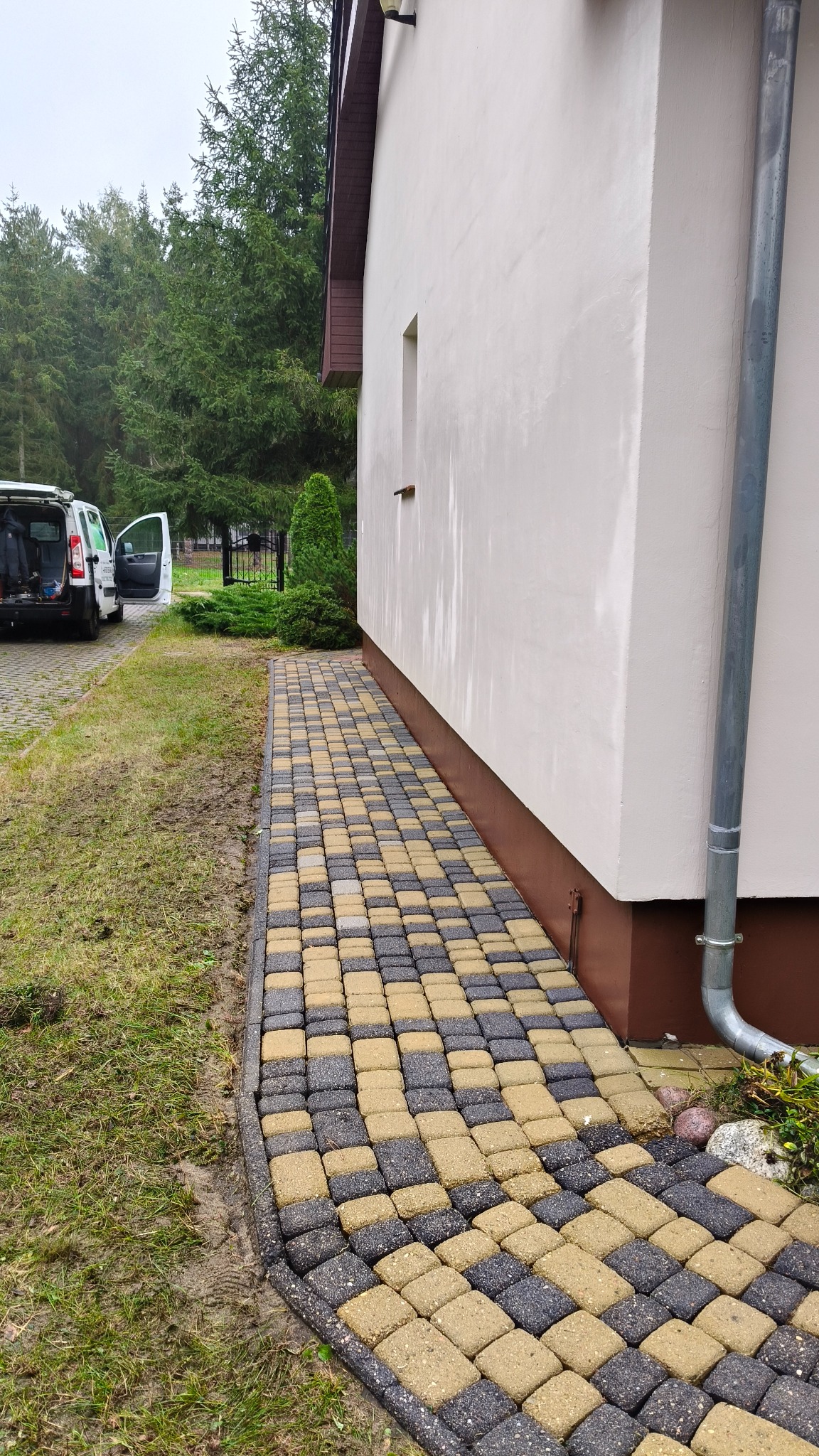 Ułożona kostka brukowa w szachownicę przy budynku w Kostrzynie nad Odrą. Widoczny fragment elewacji i rynna spustowa. W tle zieleń i zaparkowany samochód.