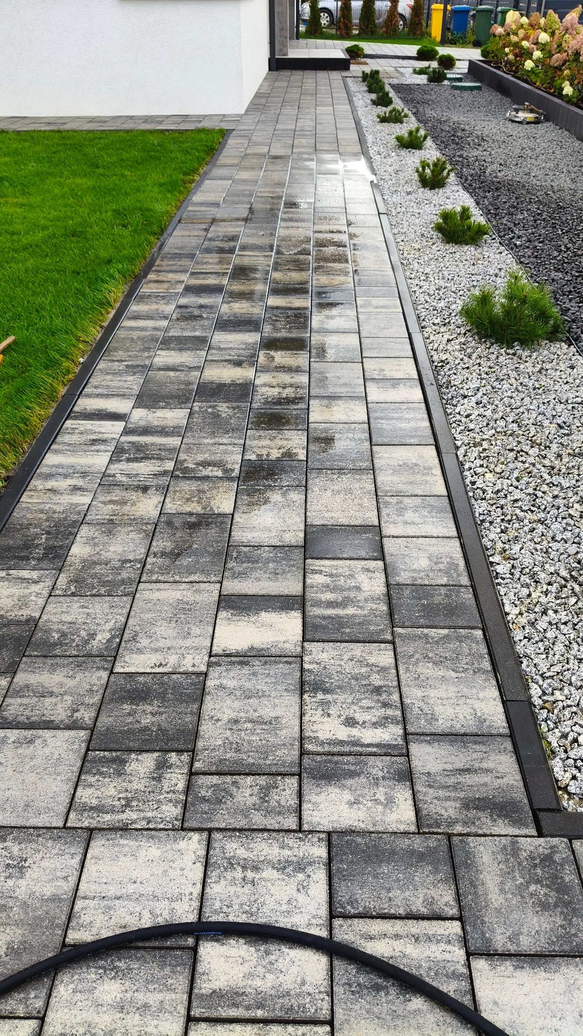 Ułożona kostka brukowa w odcieniach szarości, tworząca ścieżkę w ogrodzie. Widoczne obrzeża, trawnik i rabata z kamykami oraz roślinami. W tle budynek.
