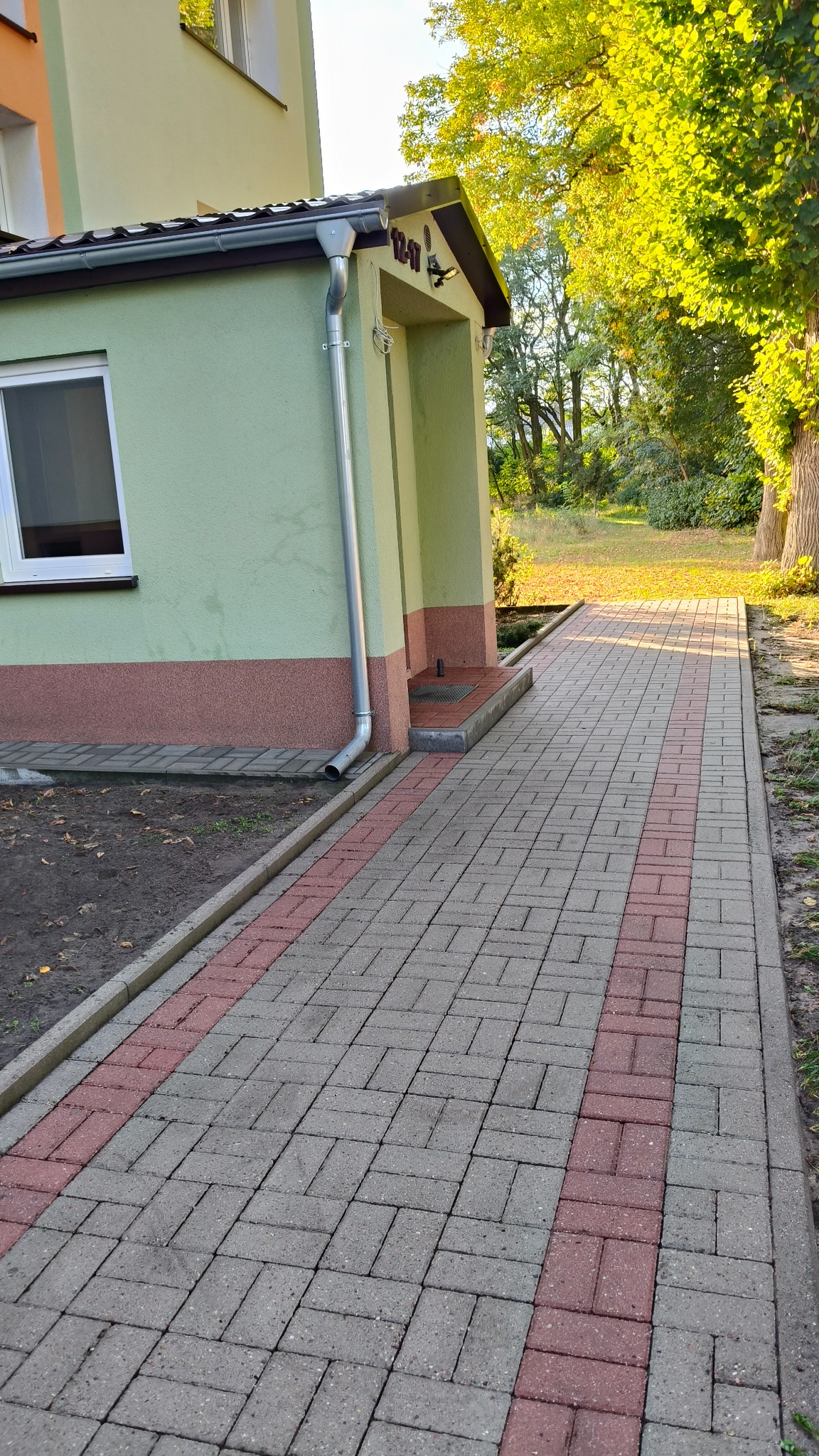 Ułożona kostka brukowa wzdłuż budynku z rynną. Szara kostka z czerwonym pasem. Widok na budynek z zieloną elewacją i fragment ogrodu. Słoneczny dzień.