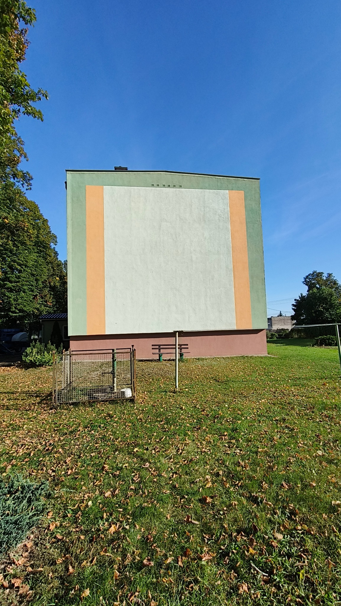 Wysoki blok z odświeżoną elewacją w pastelowych kolorach (zielony, pomarańczowy, biały). Widok z dołu na budynek otoczony trawnikiem z opadłymi liśćmi.