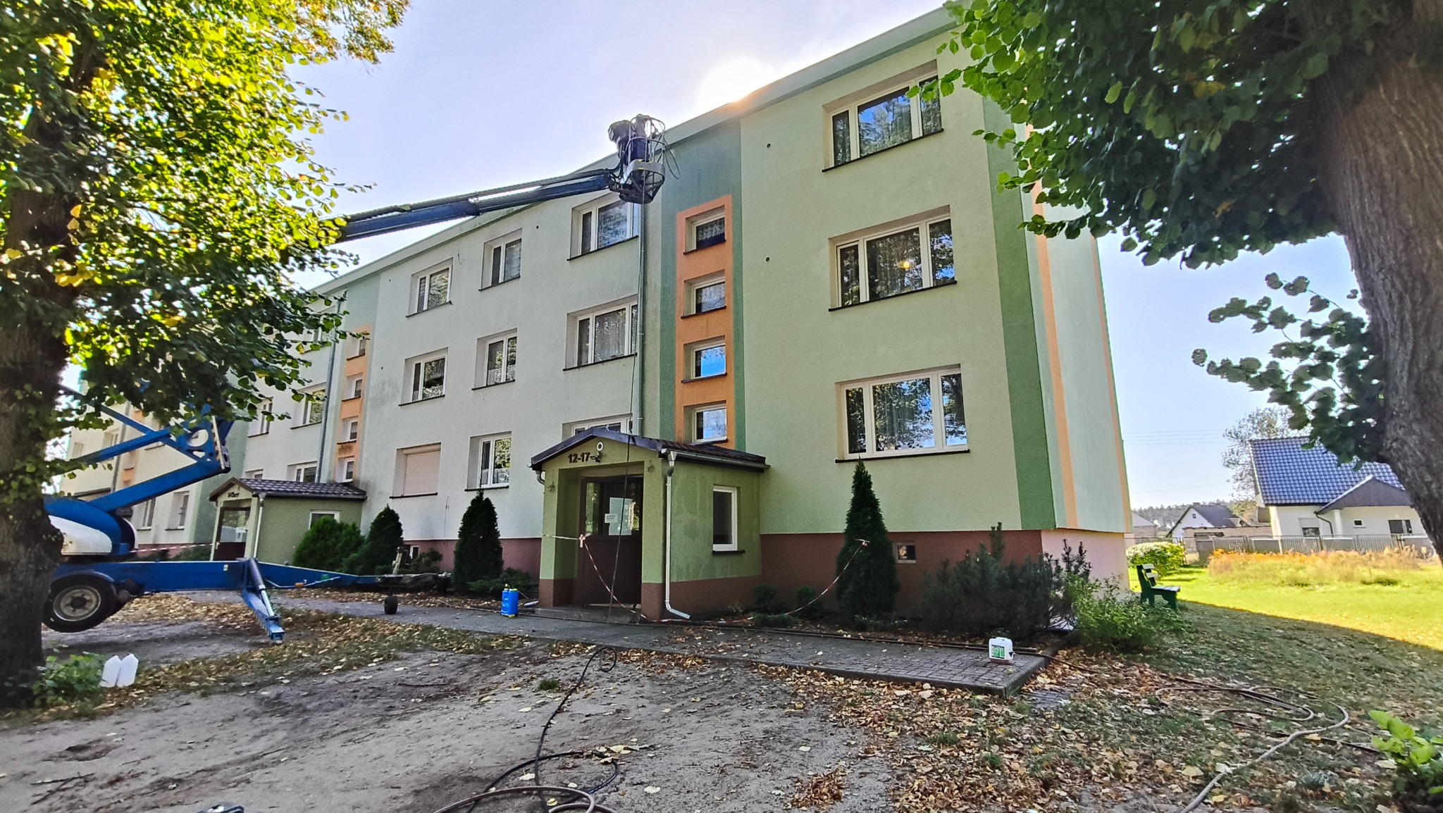 Prace na wysokościach: podnośnik koszowy przy bloku z zieloną elewacją, prawdopodobnie podczas mycia lub czyszczenia. Słoneczny dzień, widoczne drzewa.
