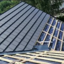 Częściowo pokryty grafitową dachówką dach z widoczną więźbą dachową i membraną w trakcie prac dekarskich.