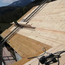 Konstrukcja drewnianego dachu w trakcie budowy, widoczna drabina transportowa i narzędzia dekarskie, w tle zalesione wzgórza.