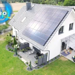 Widok z lotu ptaka na dom jednorodzinny z panelami słonecznymi na dachu, zadaszonym tarasem i zadbanym ogrodem. W tle widoczny drugi dom w stanie surowym i logo firmy SOLPRO w lewym górnym rogu.