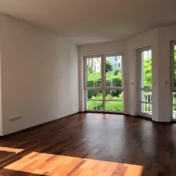 Puste jasne pomieszczenie z parkietem, trzema oknami z widokiem na zieleń i białym grzejnikiem pod jednym z nich. Na suficie widoczna instalacja elektryczna bez lampy.