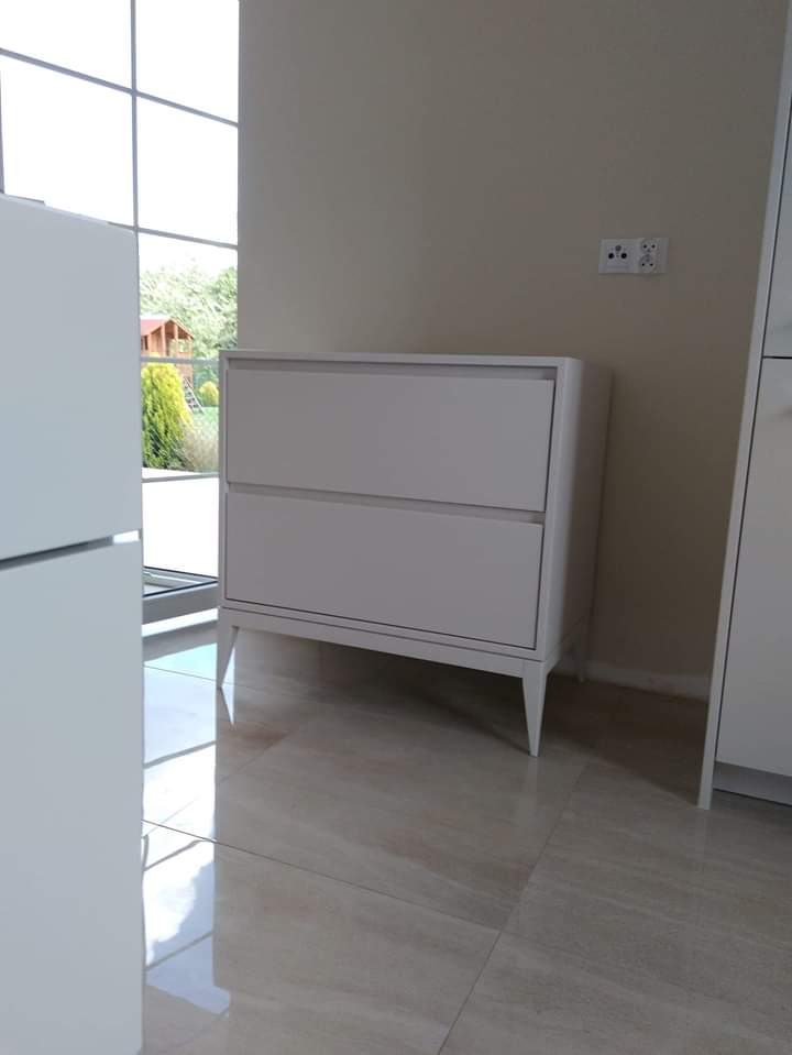 Biała, dwupoziomowa komoda na smukłych nóżkach, ustawiona w jasnym wnętrzu z płytkami podłogowymi, widok z niskiej perspektywy, okno w tle.