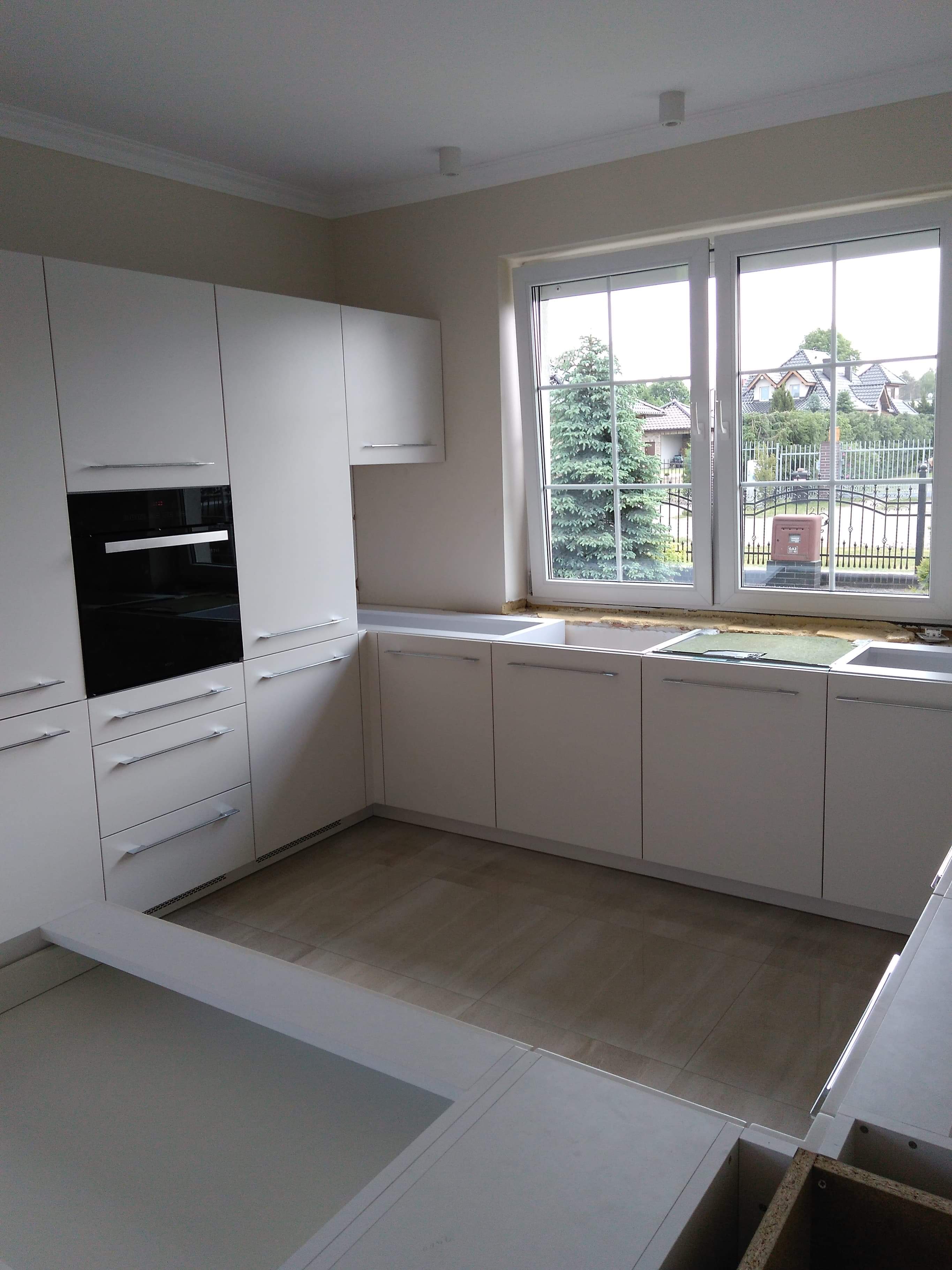 Biała, nowoczesna zabudowa kuchenna z piekarnikiem i szufladami, widok na okno z zielenią w Szczecinie, w trakcie montażu blatów.