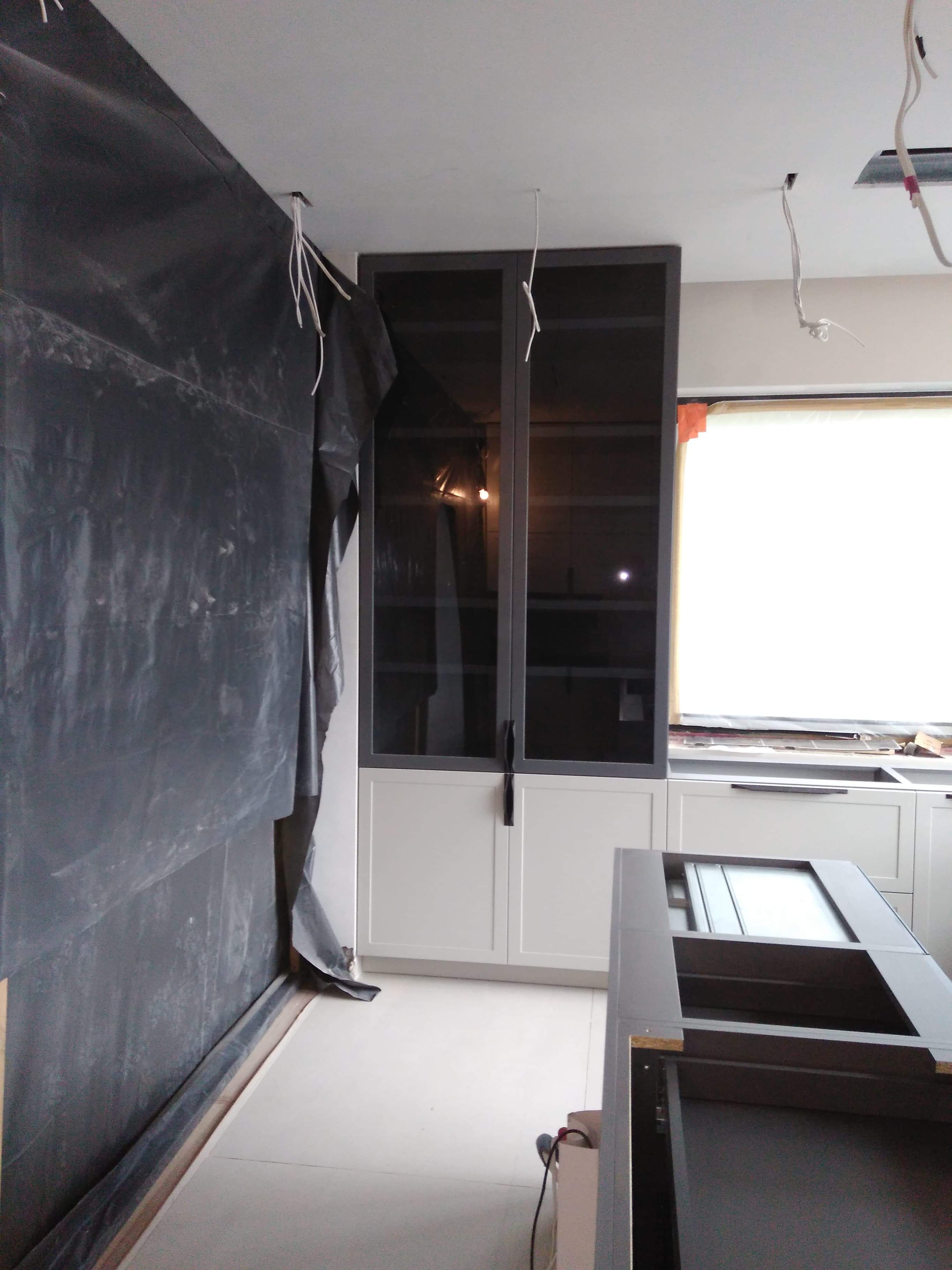 Montaż szarej szafy z przeszklonymi drzwiczkami w kuchni, zabezpieczona folią ściana i wystające kable z sufitu.