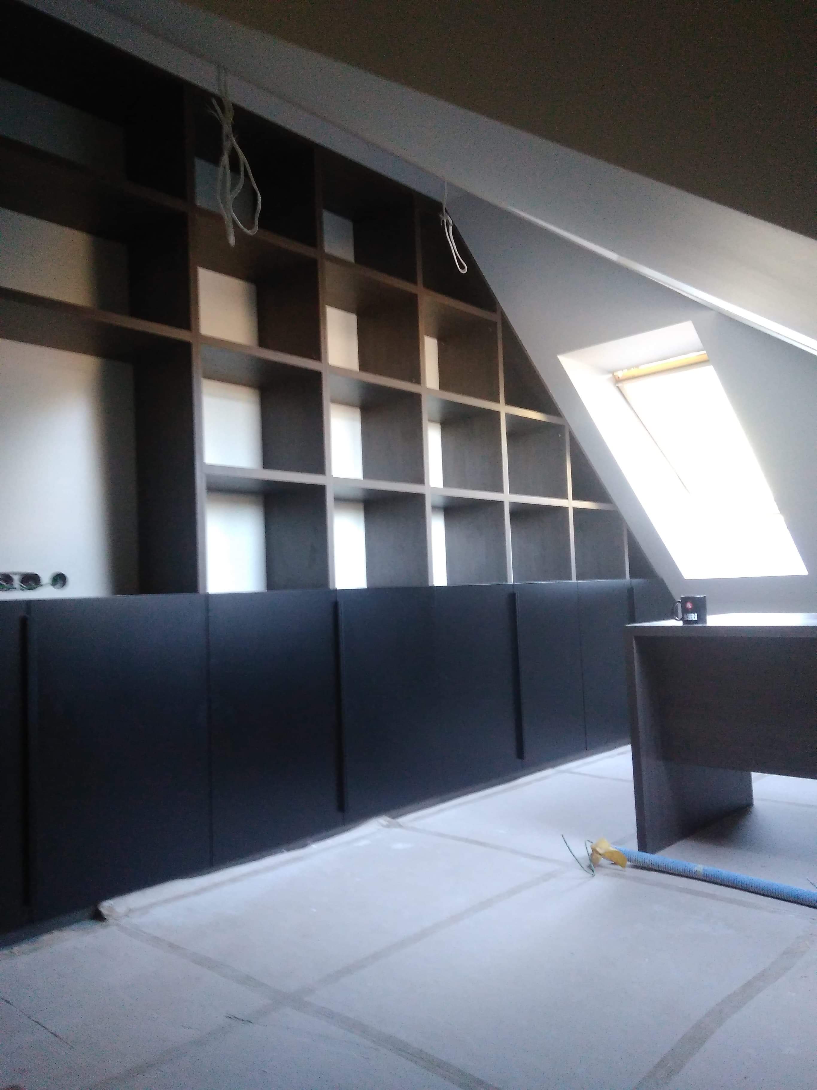 Ciemne, modułowe regały wnękowe z otwartymi półkami i dolnymi szafkami, montowane w pokoju na poddaszu ze skośnym oknem dachowym, widoczne przewody i elementy montażowe.