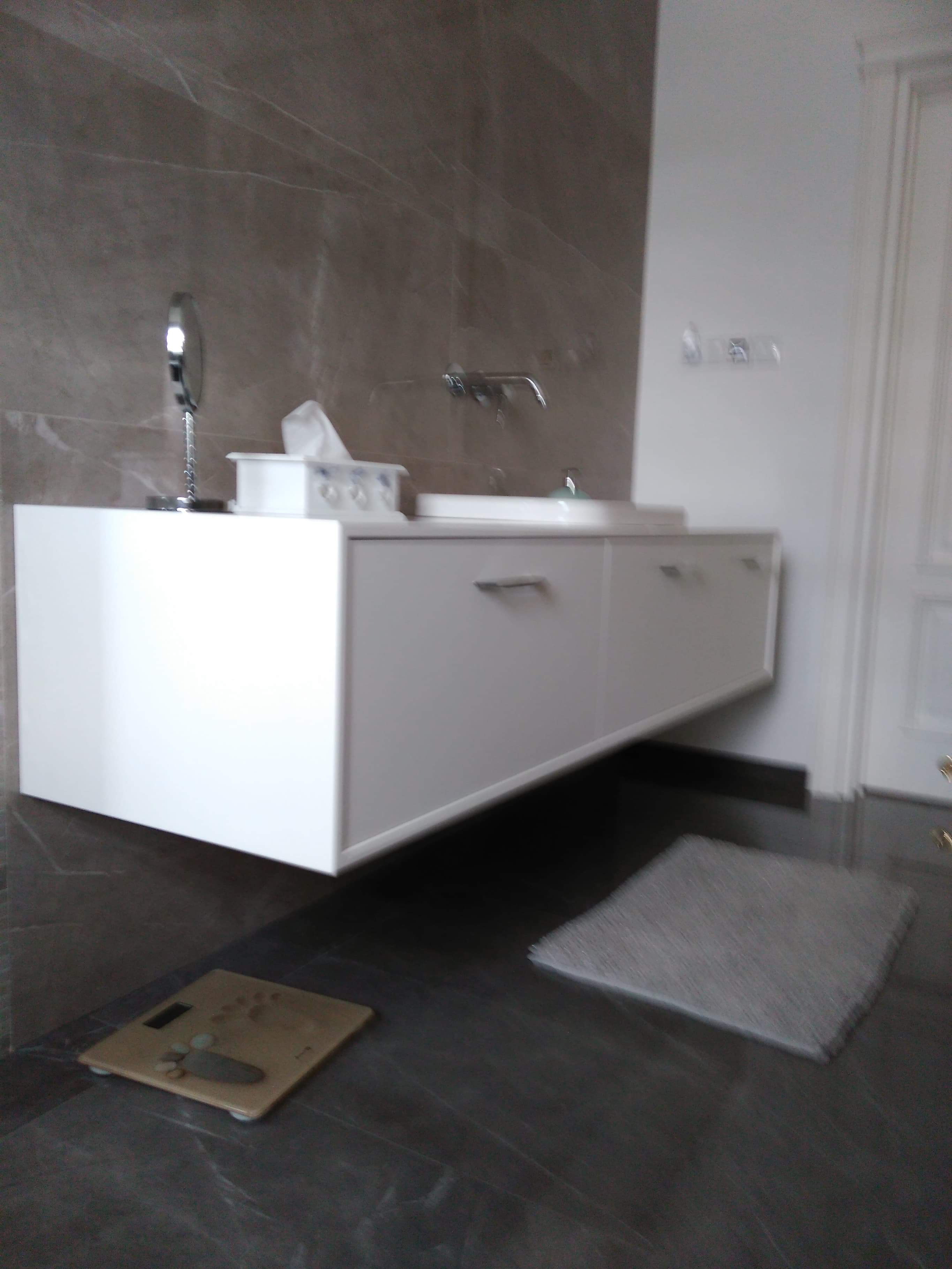 Nowoczesna, biała szafka łazienkowa pod umywalkę, zawieszona na ścianie z szarego marmuru, z widoczną baterią i akcesoriami łazienkowymi, takimi jak lusterko i pudełko na chusteczki.