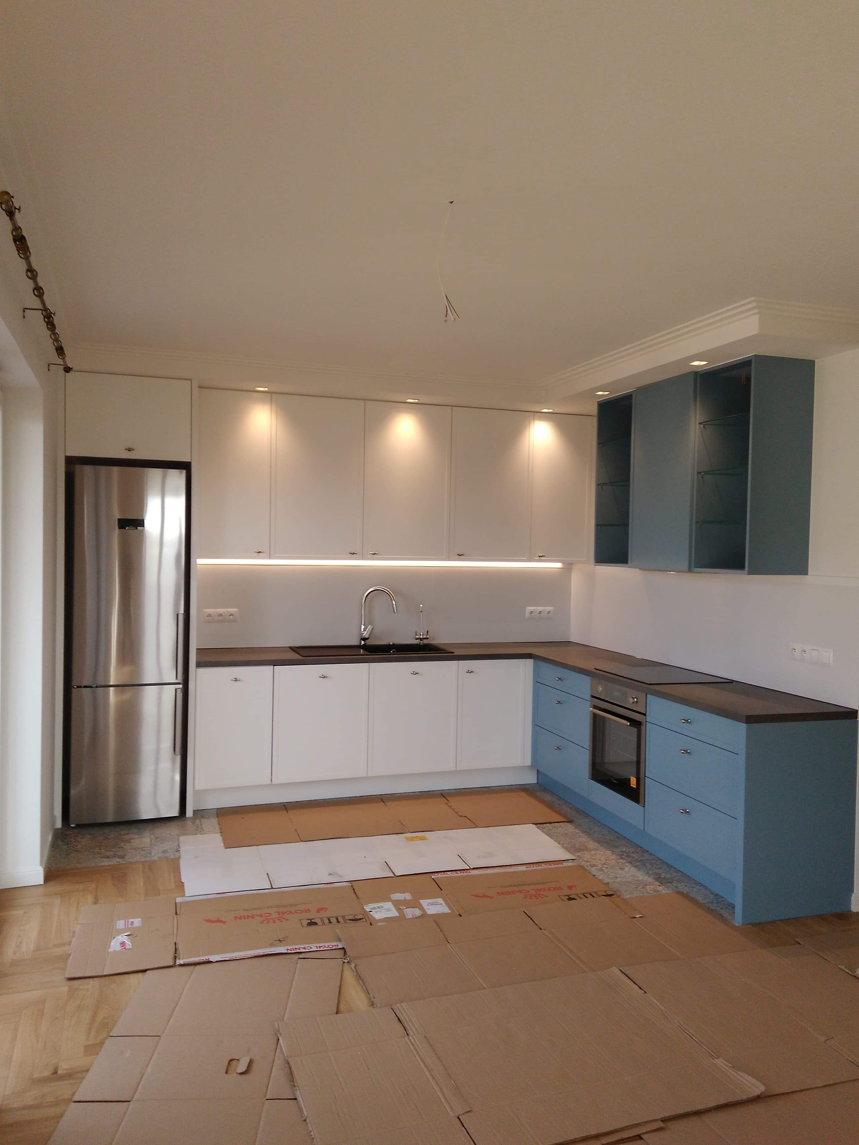 Wnętrze kuchni w trakcie montażu: białe szafki górne z oświetleniem podszafkowym, dolne szafki w odcieniach niebieskiego, stalowa lodówka i kartony zabezpieczające podłogę.