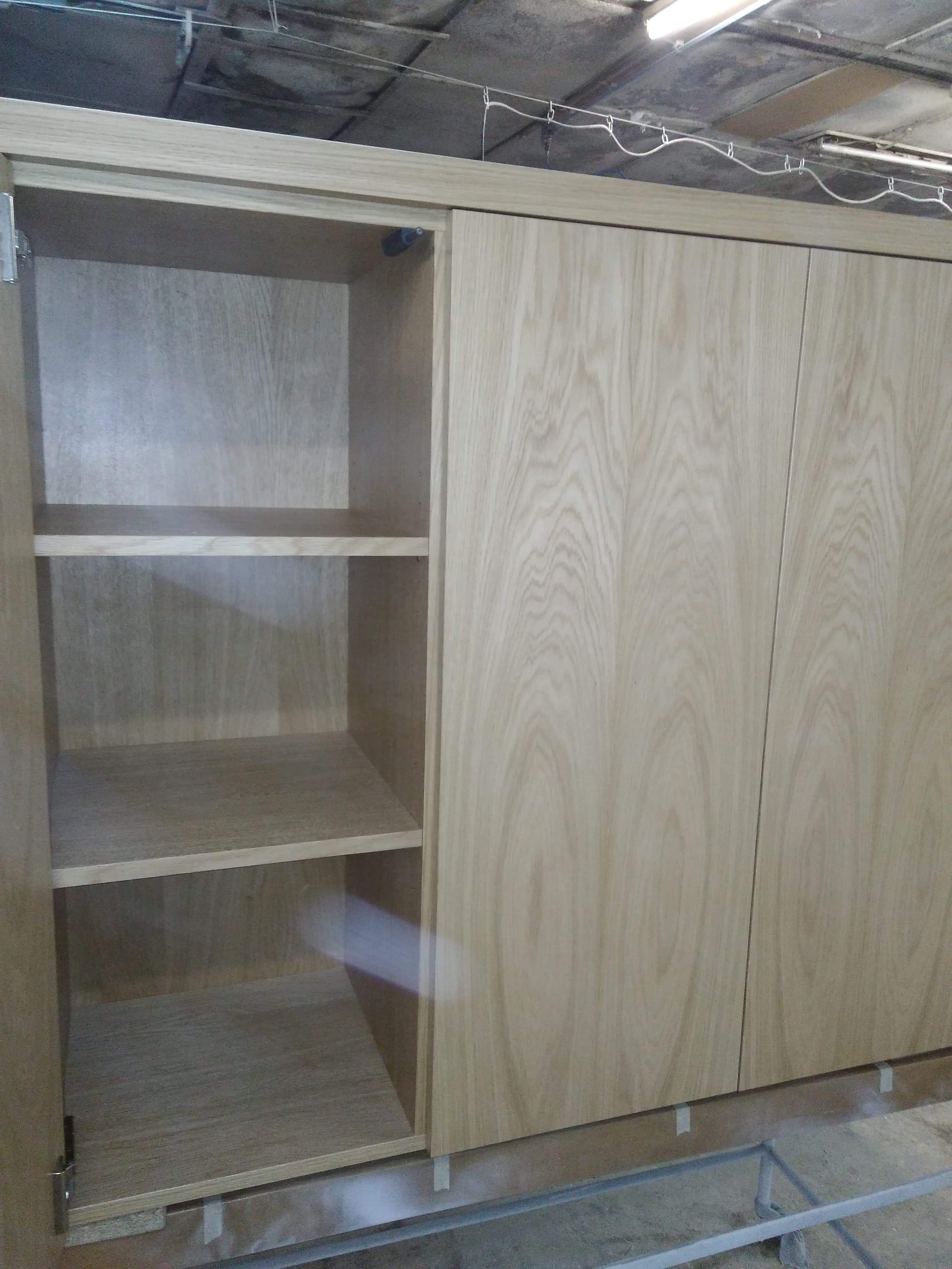 Jasna, drewniana szafa z przesuwnymi drzwiami i otwartą częścią z półkami, widoczna w warsztacie na metalowej konstrukcji.