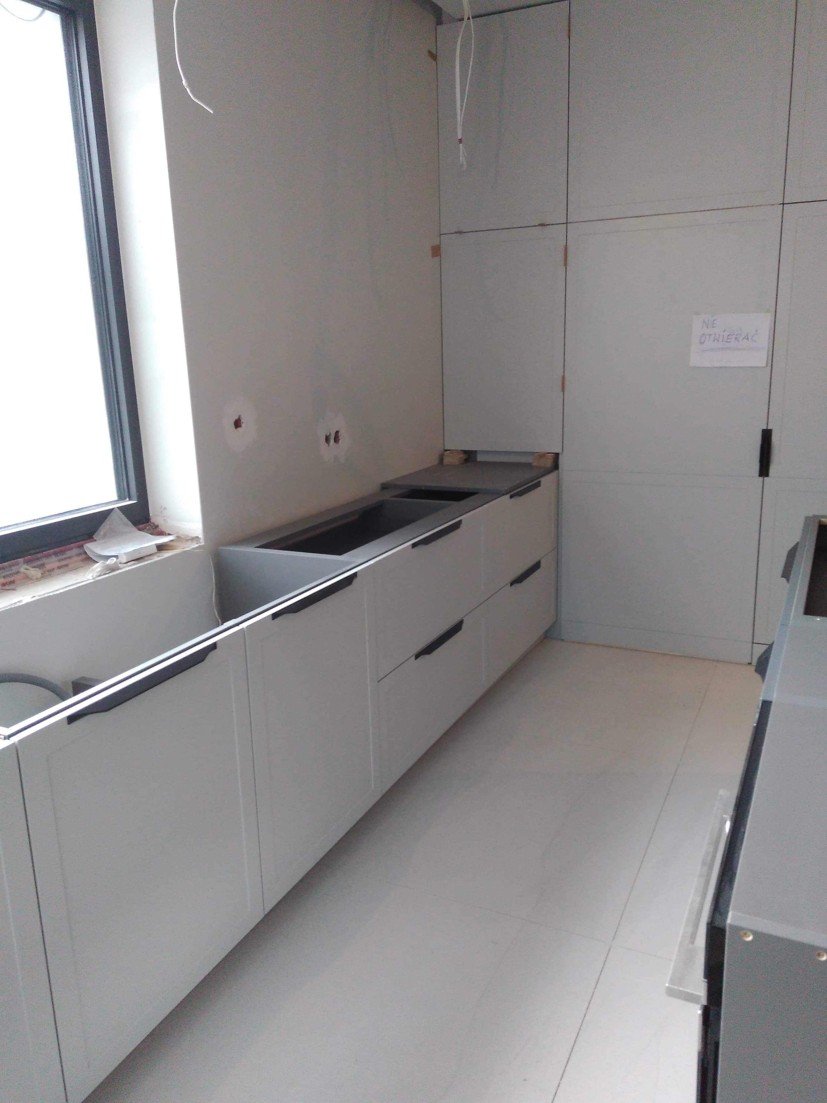 Niedokończona zabudowa kuchenna z szarymi frontami i czarnymi uchwytami, widoczne przygotowane otwory pod zlew i płytę, ściany w trakcie wykończenia, kartka z napisem 'Nie Otwierać'.