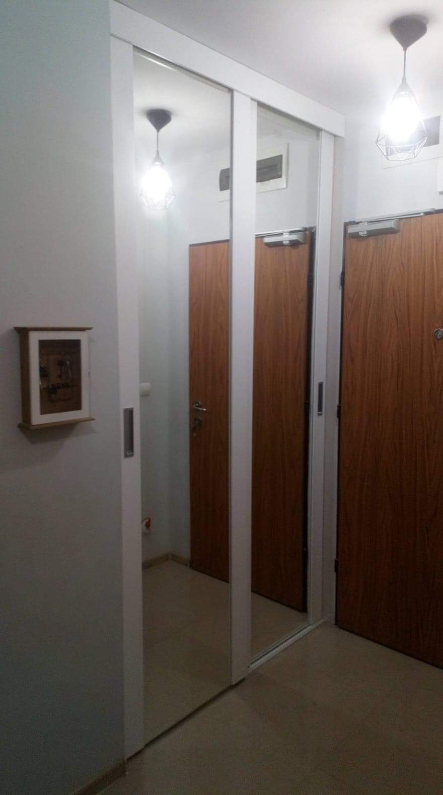 Biała szafa przesuwna z lustrzanymi drzwiami w przedpokoju, odbijająca wnętrze mieszkania, widoczne drzwi wejściowe i skrzynka na klucze na ścianie.