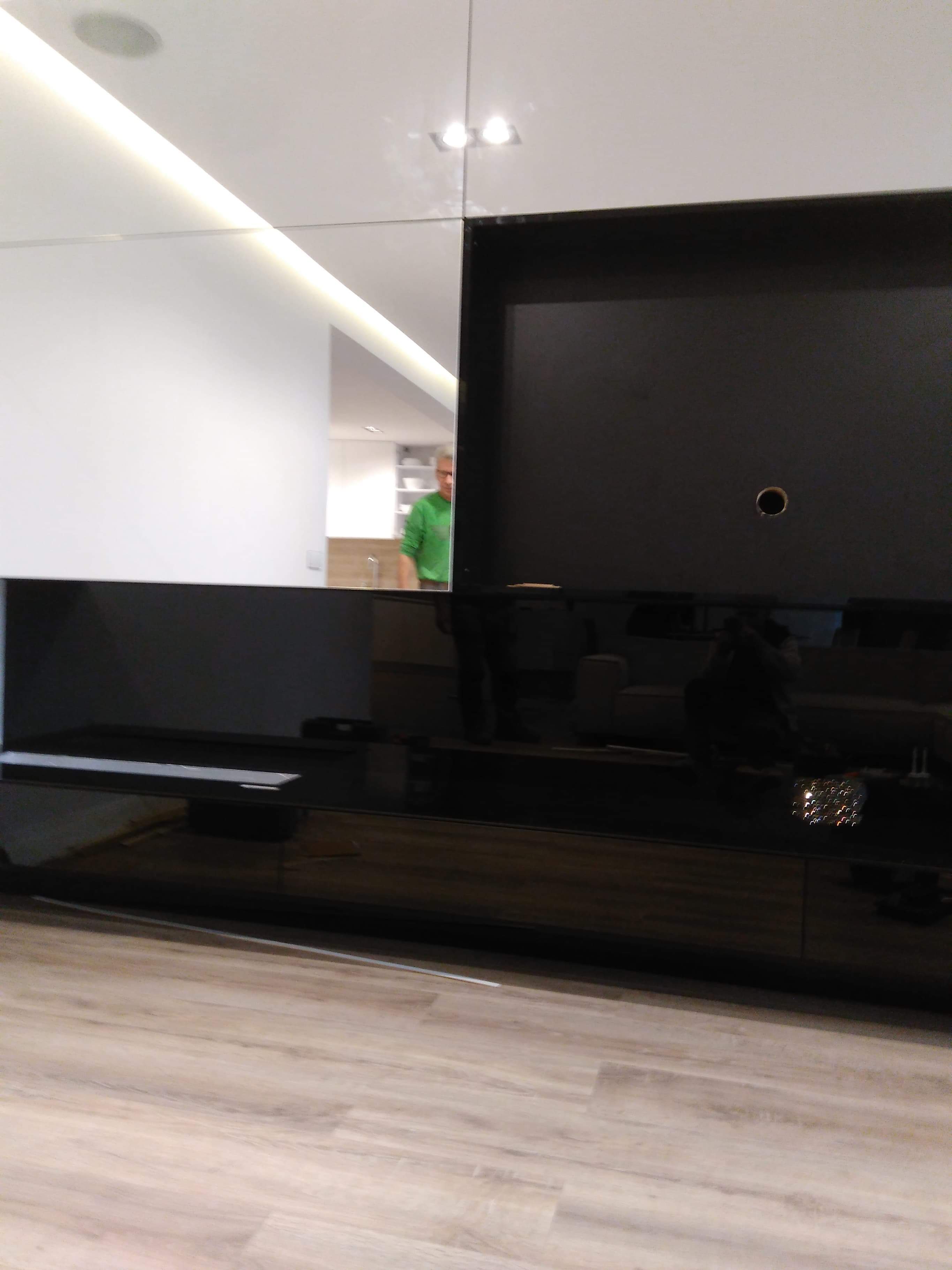 Nowoczesna szafka RTV z czarnymi i białymi frontami na wysoki połysk, widoczne odbicie osoby w kuchni.