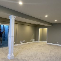 X-BUD Malowanie metodą natryskową - Wykończone wnętrze z pomalowanymi na oliwkowy kolor ścianami, jasną wykładziną i białymi listwami przypodłogowymi, widoczne kratki wentylacyjne i kolumna dekoracyjna, osoba robiąca zdjęcie w tle.