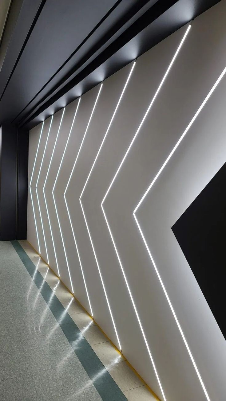 Nowoczesna ściana z białymi, ukośnymi pasami LED tworzącymi wzór strzałek, z czarnym akcentem w rogu i refleksami na podłodze.