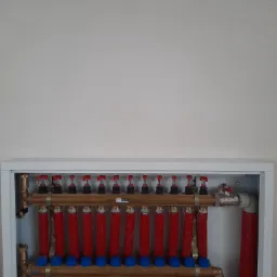 Rozdzielacz podłogowy z czerwonymi rurami w białej szafce, widoczne zawory i przyłącza mosiężne.