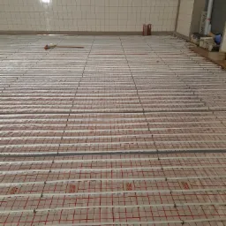 Rozłożona instalacja ogrzewania podłogowego z rurkami na folii z czerwoną kratką w pomieszczeniu z białymi płytkami na ścianach. W tle widoczne rury i materiały budowlane.