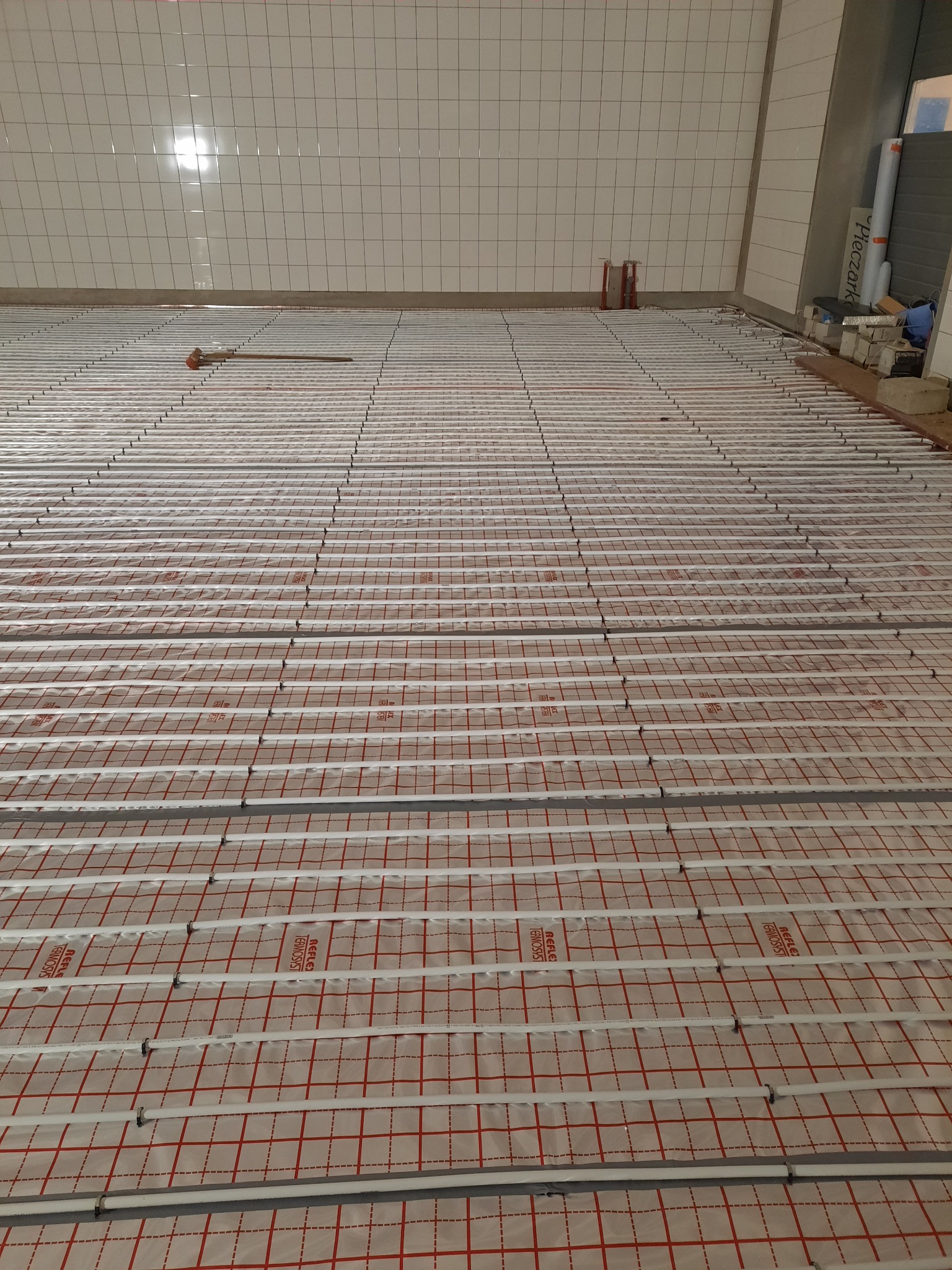Rozłożona instalacja ogrzewania podłogowego z rurkami na folii z czerwoną kratką w pomieszczeniu z białymi płytkami na ścianach. W tle widoczne rury i materiały budowlane.