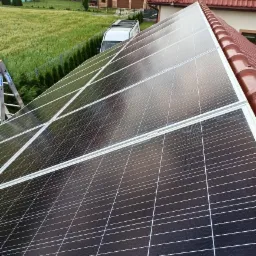 Panele fotowoltaiczne zamontowane na dachu domu w Bezwoli, widok z góry, drabina oparta o dach, w tle zabudowa jednorodzinna i bus firmy.