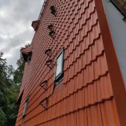 Czerwona dachówka ceramiczna na stromym dachu domu z oknami dachowymi i wspornikami drabiny dachowej, widok z dołu pod kątem w pochmurny dzień.