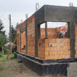 Wznoszenie ścian parterowego domu z czerwonej ceramiki poryzowanej. Widoczne zbrojenia pionowe i betonowe nadproża. W tle drzewa i pranie na sznurze.