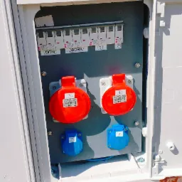 Rozdzielnica elektryczna z bezpiecznikami i gniazdami siłowymi, zamontowana na ceglanej ścianie budynku. Widoczne oznaczenia 'DOM' i 'OBORA' na bezpiecznikach.