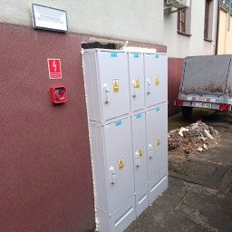 ELEKTROTECHNIKA MICHAŁ KASZUBA - Zewnętrzna szafa elektryczna, zabezpieczona pianką montażową, z widoczną skrzynką przeciwpożarową i oświetleniem elewacyjnym na ścianie budynku.