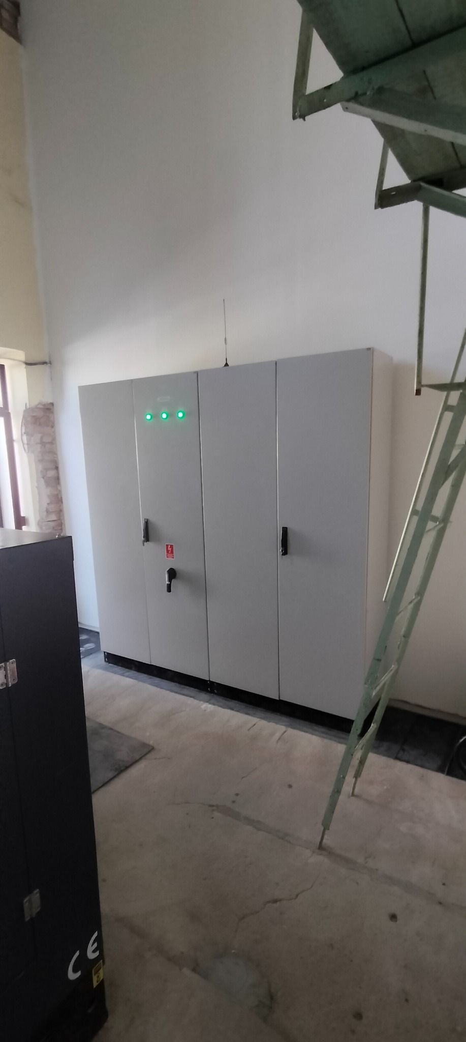 Szara szafa elektryczna z trzema zielonymi diodami i anteną na górze, zamontowana na ścianie w surowym wnętrzu. Widoczna drabina i fragment urządzenia.