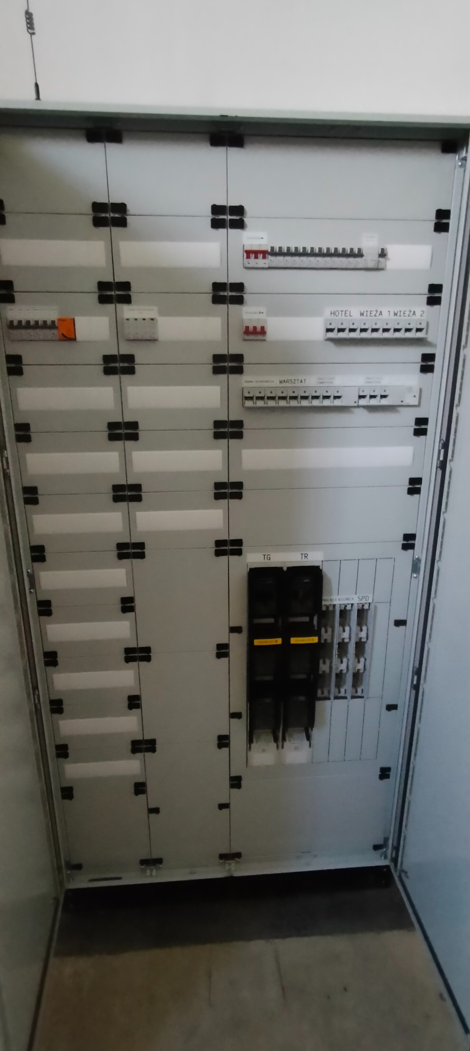 Otwarta skrzynka elektryczna z bezpiecznikami i opisami 'Hotel Wieża 1 Wieża 2' i 'Warsztat'. Widoczne zabezpieczenia i listwy zaciskowe. Szara obudowa.