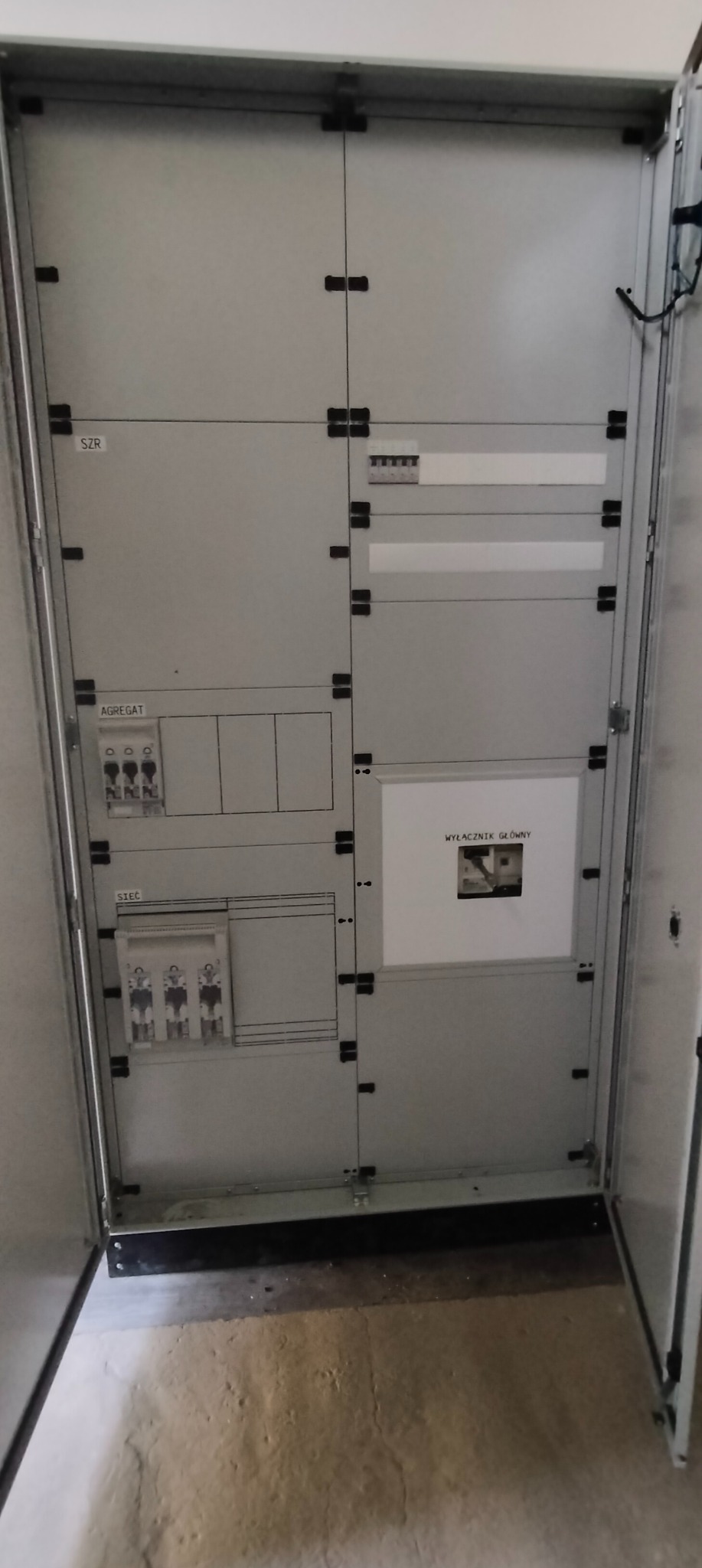 Otwarta, szara szafa elektryczna z opisami 'SZR', 'Agregat', 'Sieć', 'Wyłącznik główny' oraz widocznymi bezpiecznikami i elementami instalacji.