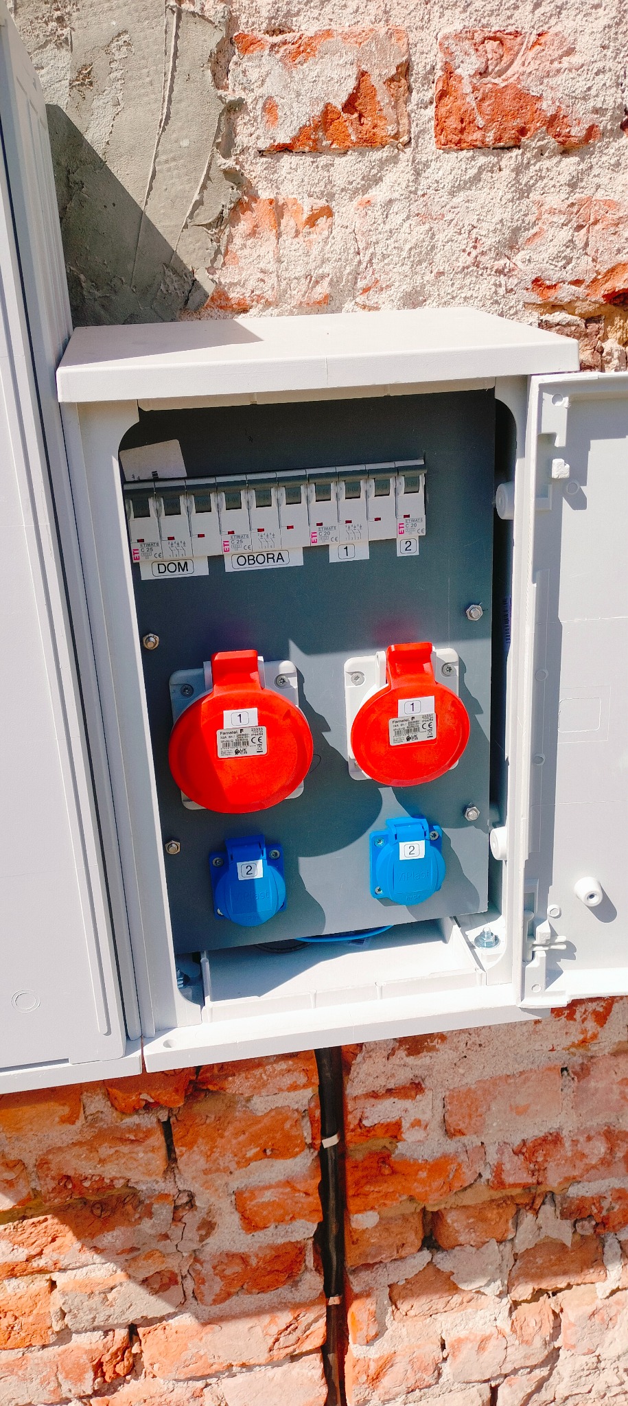 Rozdzielnica elektryczna z bezpiecznikami i gniazdami siłowymi, zamontowana na ceglanej ścianie budynku. Widoczne oznaczenia 'DOM' i 'OBORA' na bezpiecznikach.