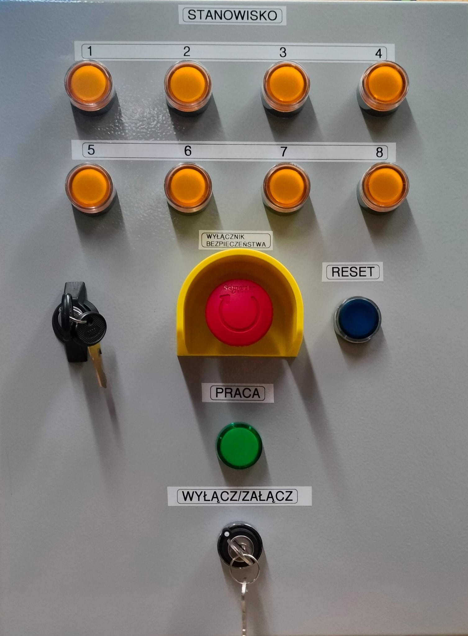 Szara skrzynka sterownicza z ośmioma pomarańczowymi przyciskami, wyłącznikiem bezpieczeństwa, przyciskiem reset, przyciskiem praca i dwoma kluczami w stacyjce.