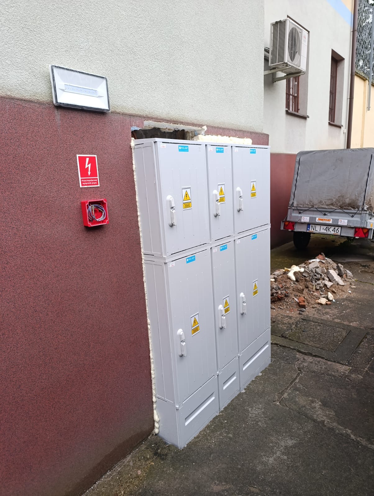 Zewnętrzna szafa elektryczna, zabezpieczona pianką montażową, z widoczną skrzynką przeciwpożarową i oświetleniem elewacyjnym na ścianie budynku.