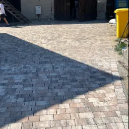 Nowo ułożona, wielobarwna kostka brukowa na podjeździe z widocznym cieniem oraz fragmentem budynku gospodarczego i żółtego pojemnika na śmieci w tle.