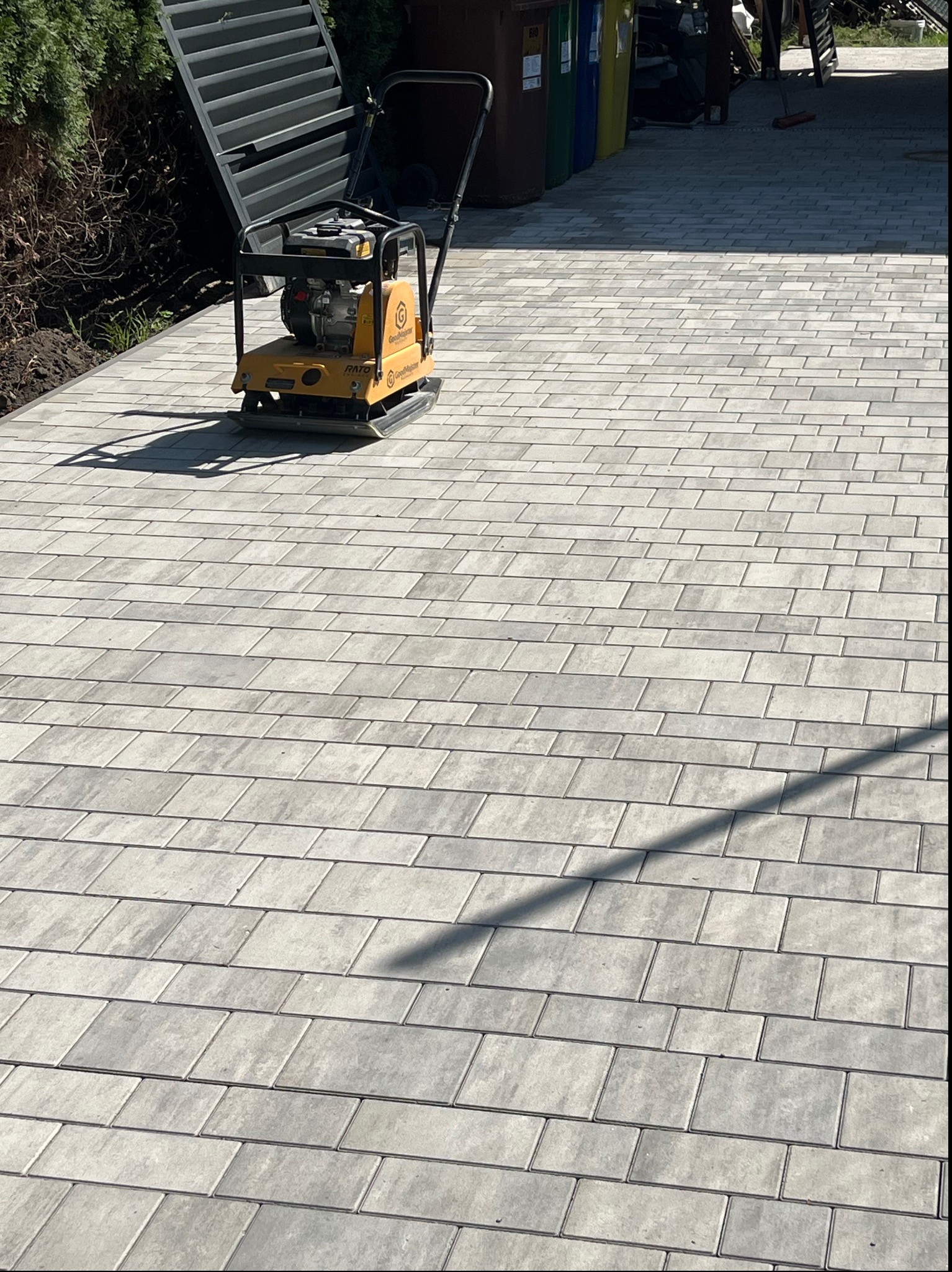 Żółta zagęszczarka obok świeżo ułożonej, szarej kostki brukowej, widoczne ślady pracy i cień urządzenia.