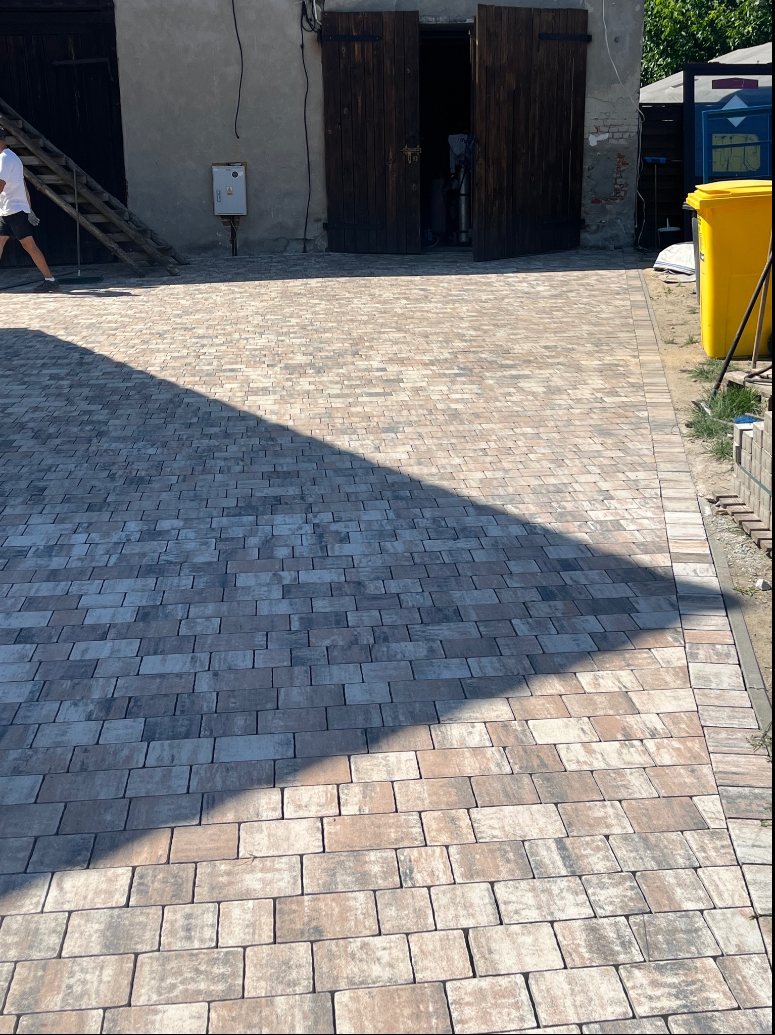Nowo ułożona, wielobarwna kostka brukowa na podjeździe z widocznym cieniem oraz fragmentem budynku gospodarczego i żółtego pojemnika na śmieci w tle.