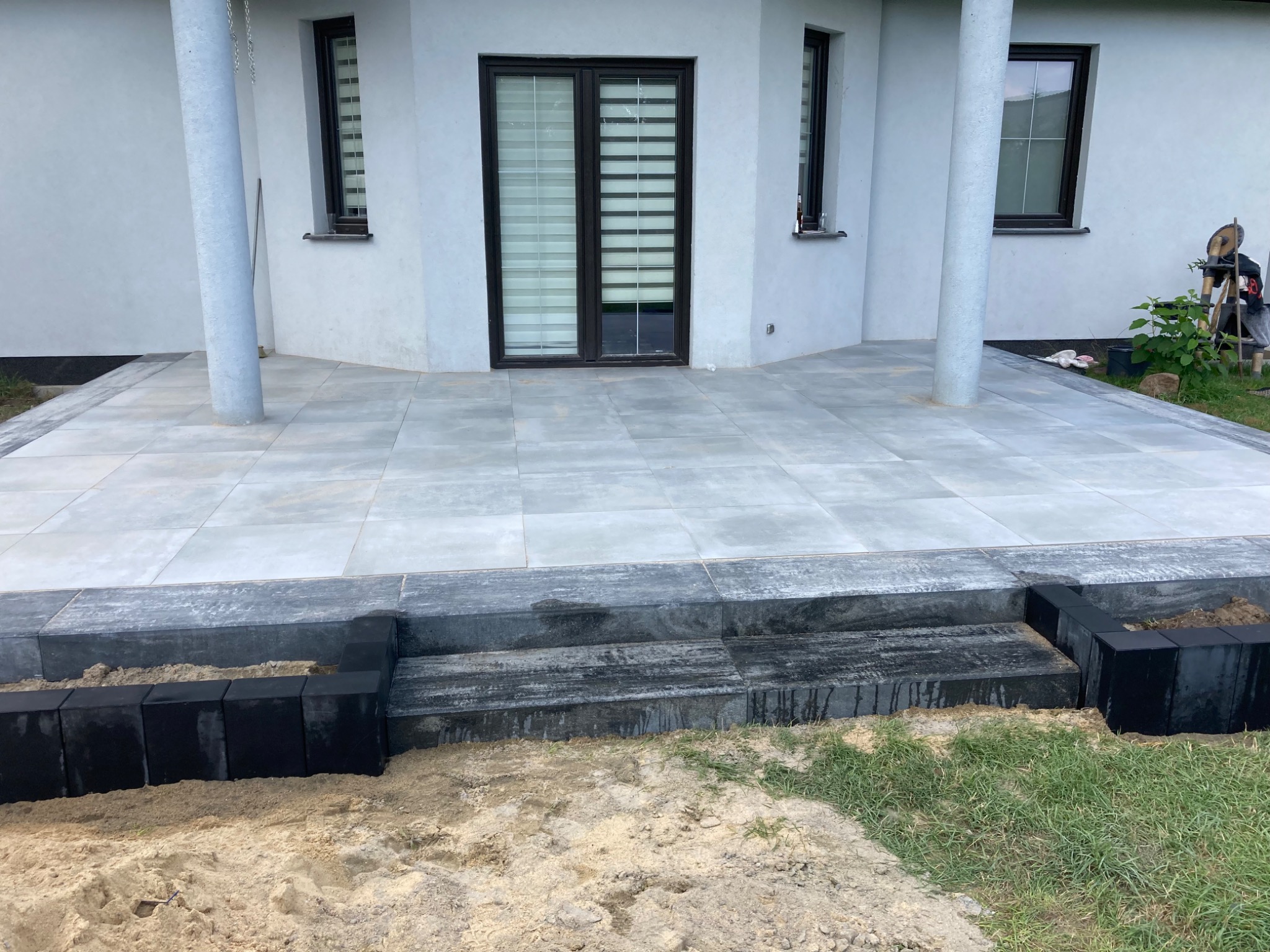 Nowoczesny taras z szarych płyt betonowych, z czarnymi obrzeżami i schodami, widok na biały dom z oknami i kolumnami, piasek i trawa w tle.