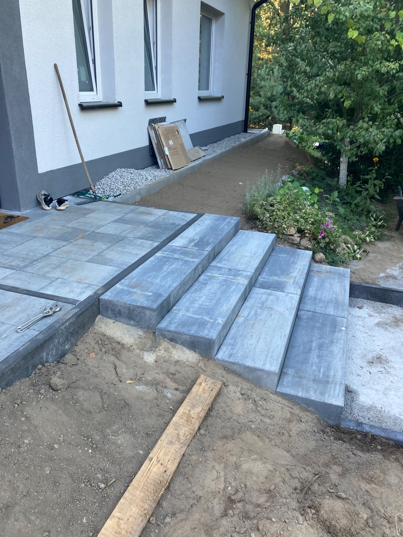 Schody zewnętrzne z szarych, betonowych elementów w trakcie budowy, widoczne narzędzia i ziemia, dom z białymi ścianami w tle.