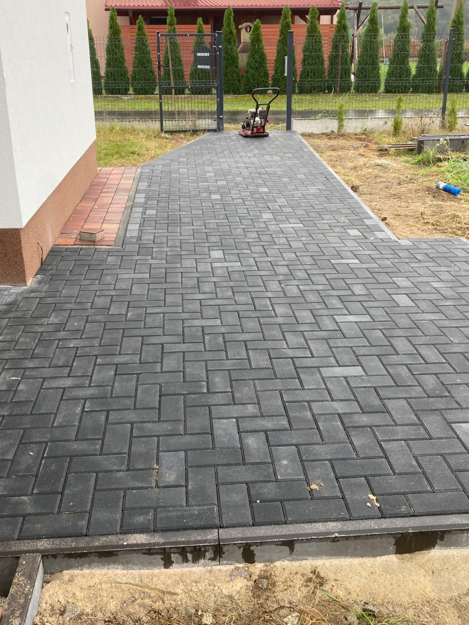 Ułożona, grafitowa kostka brukowa w jodełkę prowadząca od domu do metalowej bramki z widoczną zagęszczarką w tle.