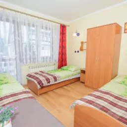 Jasny pokój z trzema pojedynczymi łóżkami, szafą i oknem z czerwonymi i białymi zasłonami. Na jednym z łóżek stoi doniczka z fioletowymi kwiatami.