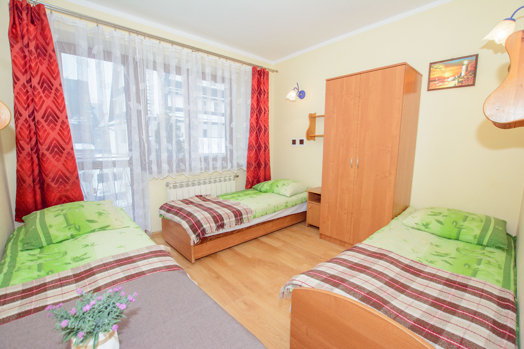 Jasny pokój z trzema pojedynczymi łóżkami, szafą i oknem z czerwonymi i białymi zasłonami. Na jednym z łóżek stoi doniczka z fioletowymi kwiatami.